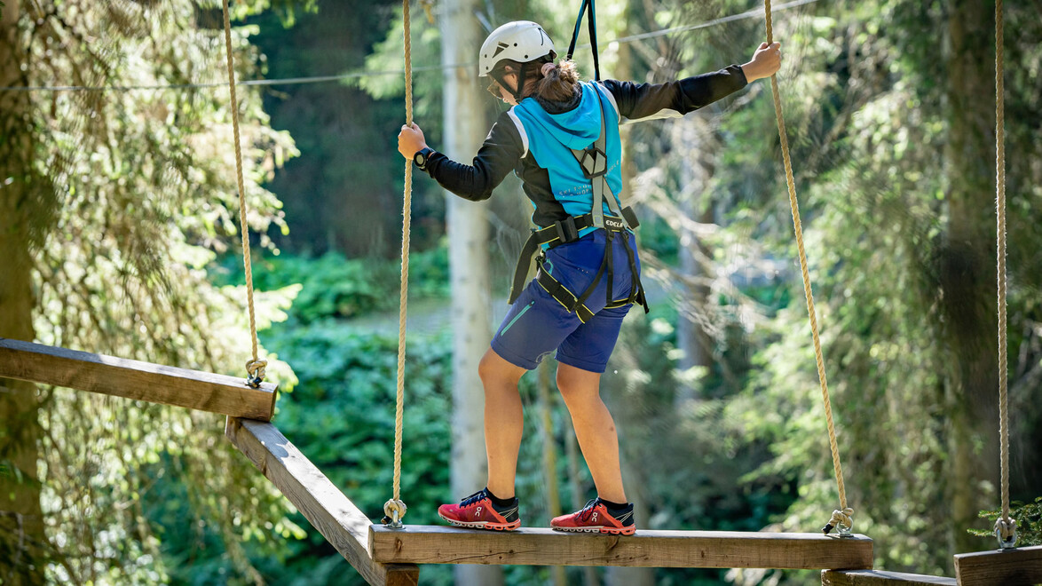 Mann balanciert auf schmalem Balken im Hochseilgarten, gesichert mit Seil / Man balances on narrow beam in high ropes course, secured with rope