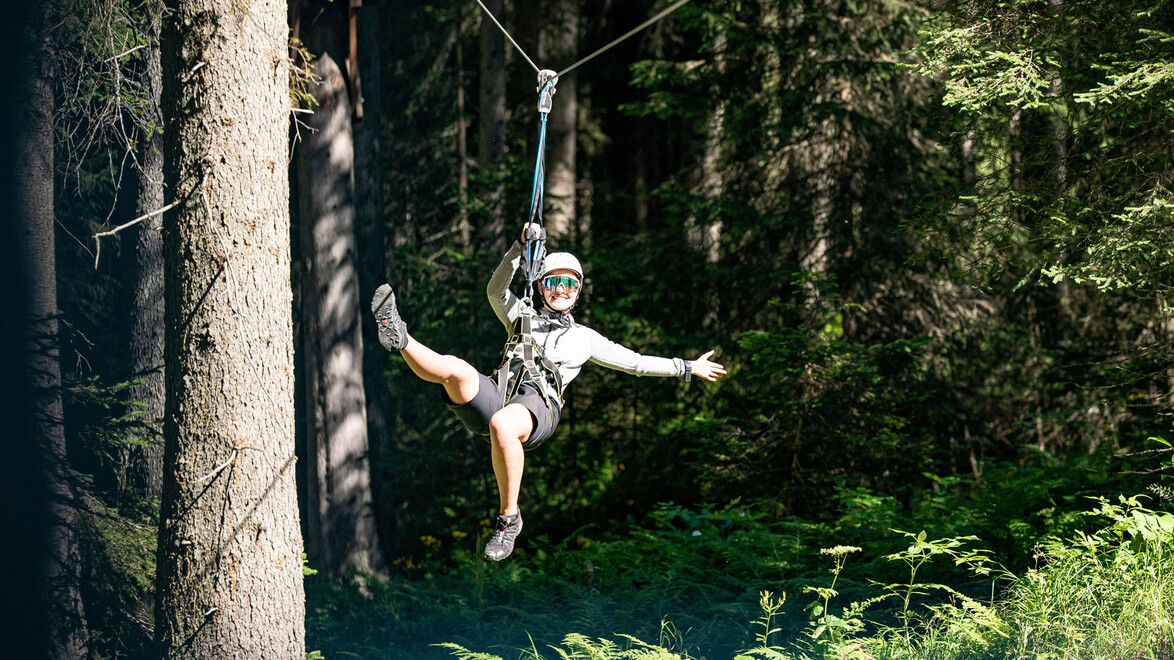 Person in grauem Outfit fährt mit einem Seil über den Wald, Beine ausgestreckt / Person in grey outfit glides over the forest on a rope, legs extended