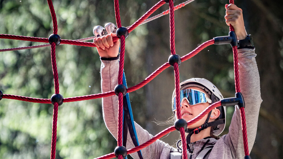 Kind mit Helm greift ein rotes Kletternetz im Hochseilgarten, konzentrierter Blick / Child with helmet grips red climbing net in high ropes course, focused expression