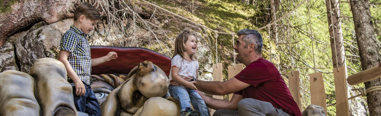 Two kids and a man laugh at a forest legend station with a troll figure, surrounded by roots and wooden elements.