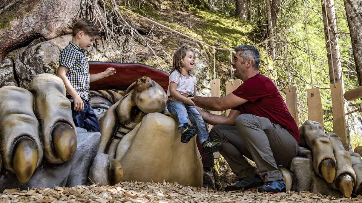 Two kids and a man laugh at a forest legend station with a troll figure, surrounded by roots and wooden elements.