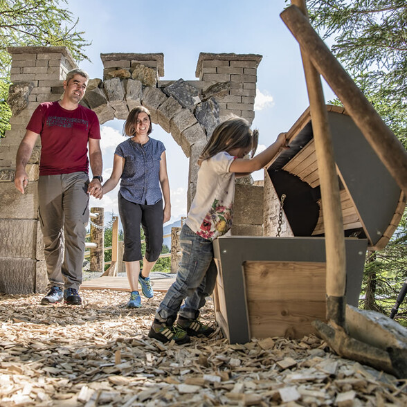 A child opens a wooden chest in the forest while parents smile nearby, next to a playground with a castle wall.