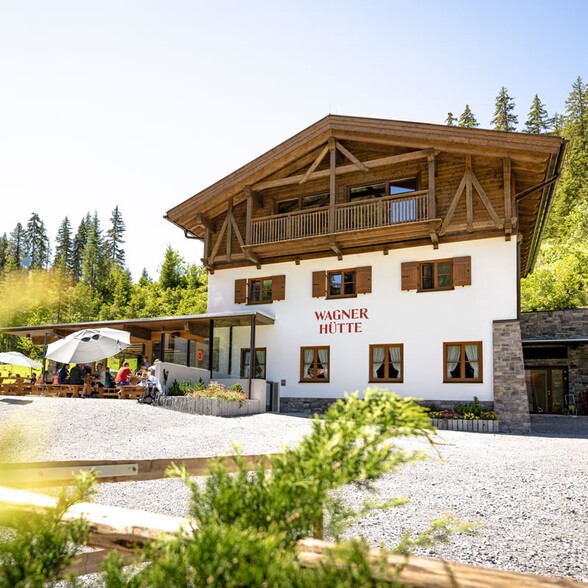 Wagner Hütte sits sunny by the forest, guests relax on the terrace, surrounded by alpine nature and mountains.