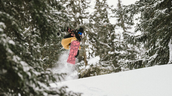 Snowboarder jumping into deep snow between snow-covered trees.