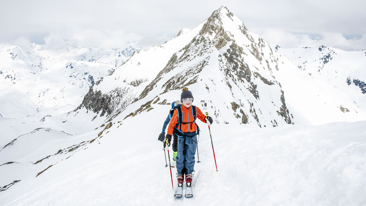 Ein Skitourengeher mit orangefarbener Jacke steht auf einem verschneiten Grat vor einem markanten Berggipfel / A ski tourer in an orange jacket stands on a snowy ridge facing a prominent mountain peak