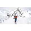 Ein Skitourengeher mit orangefarbener Jacke steht auf einem verschneiten Grat vor einem markanten Berggipfel / A ski tourer in an orange jacket stands on a snowy ridge facing a prominent mountain peak