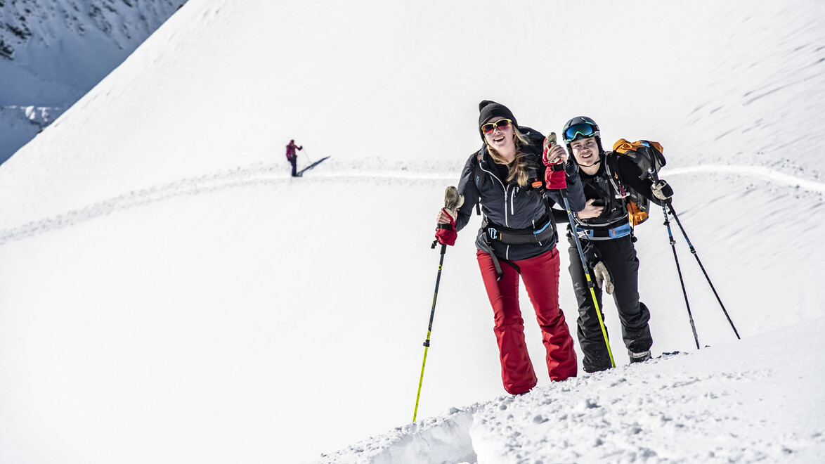 Zwei Skitourengeherinnen gehen nebeneinander eine steile Schneefläche hinauf, im Hintergrund weitere Tourengeher / Two ski tourers hike up a steep snowfield side by side, with others in the background