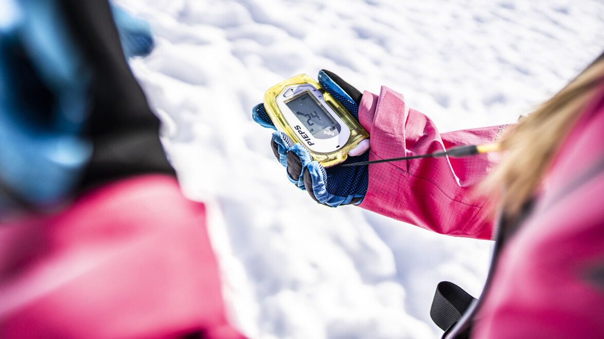Detailaufnahme eines LVS-Geräts in einer behandschuhten Hand auf dem Schnee zur Lawinensicherheit / Close-up of an avalanche transceiver in a gloved hand on the snow for avalanche safety