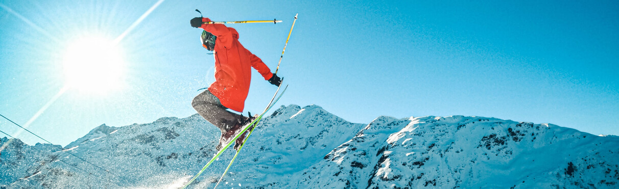 Freestyle skier jumps into the air in front of snowy Alpine scenery and bright sunshine.