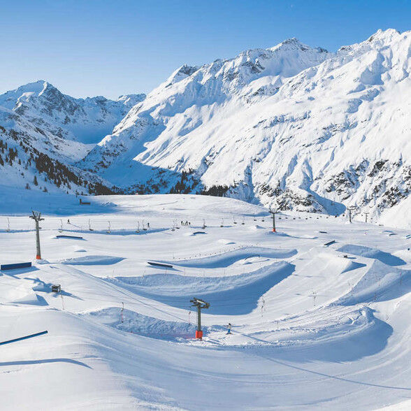 Overview of the snow park with rails, jumps, and snowy mountain peaks in the background.