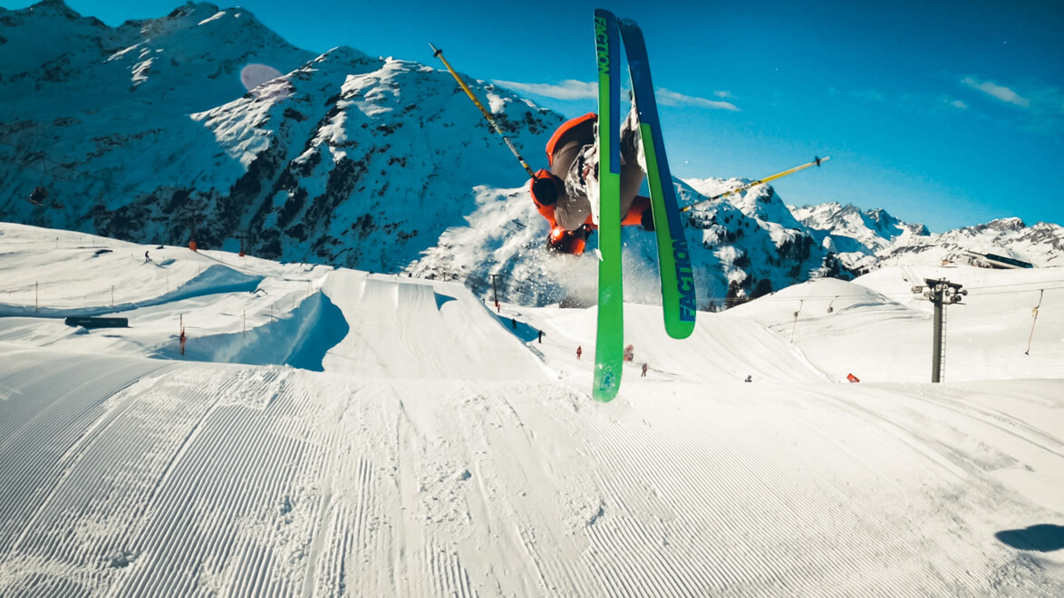 Freeskier fliegt mit überkreuzten Skiern über eine große Schanze vor hochalpiner Kulisse / Freeskier crosses skis in mid-air over a large jump with a high alpine backdrop