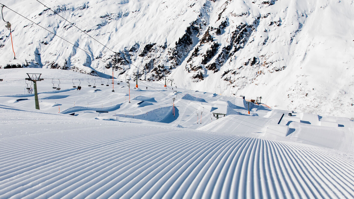 Perfekt präparierte Piste mit Blick auf den Snowpark und umliegende Berge bei Sonnenschein / Perfectly groomed slope with a view of the snow park and surrounding mountains in sunshine