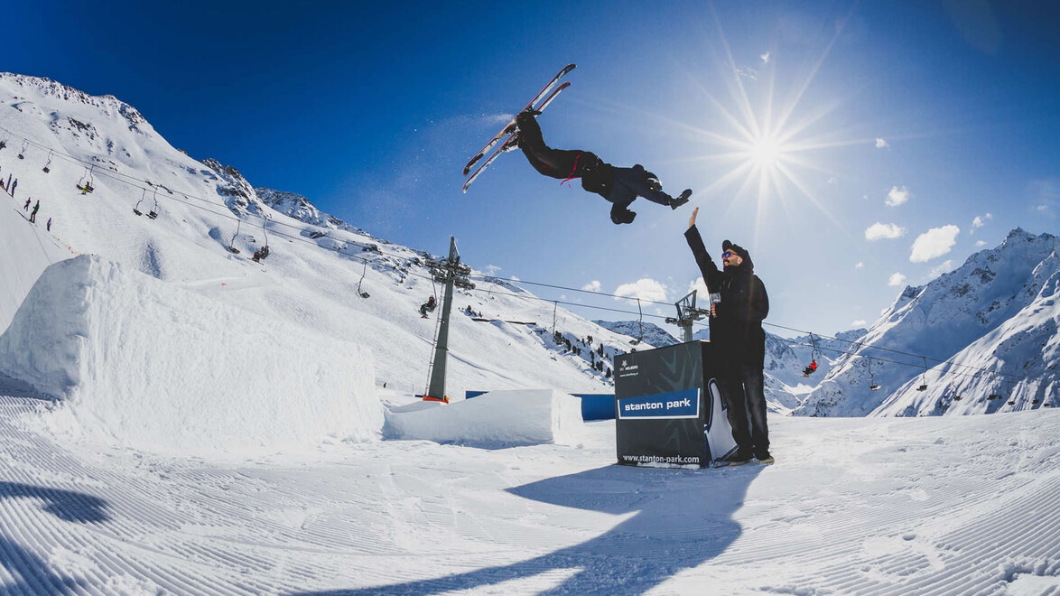 Snowboarder macht einen Sprung vor Publikum im Snowpark, während die Sonne tief steht / Snowboarder performs a jump in front of spectators in the snow park under a low sun