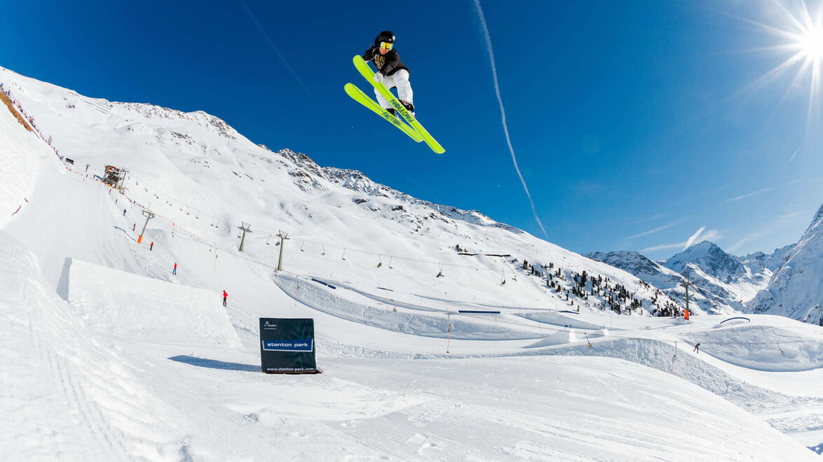 Ein Skifahrer mit neongrünen Ski springt über eine Schneerampe im Snowpark vor blauen Himmel / A skier with neon green skis jumps over a snow ramp in the snow park under blue sky