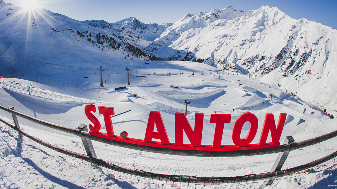 Rote Buchstaben mit „St. Anton“ stehen vor verschneiter Bergkulisse mit Sessellift im Hintergrund / Red “St. Anton” letters in front of a snowy mountain backdrop with a chairlift in the background