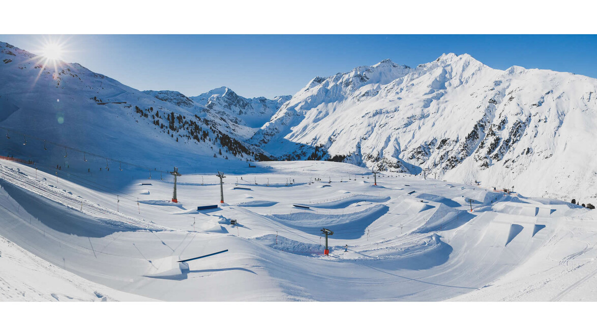 Panoramaaufnahme des Snowparks mit mehreren Schanzen und Rails, umgeben von verschneiten Bergen / Panoramic view of the snow park with jumps and rails, surrounded by snowy mountains