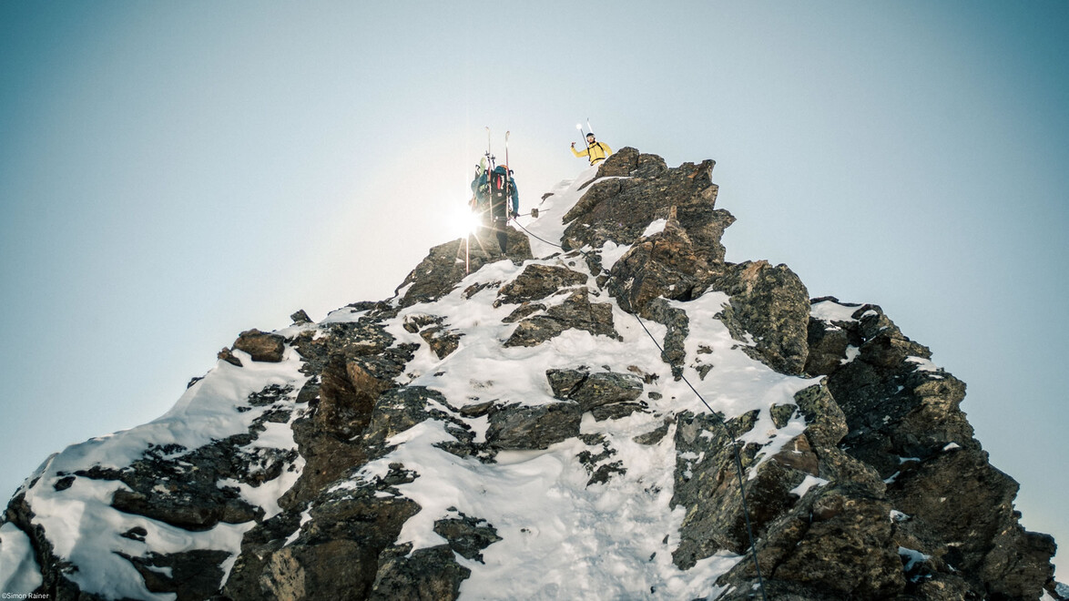 Zwei Personen erreichen den felsigen Gipfel einer Wintertour, Sonnenlicht scheint über den Grat / Two people reach the rocky summit of a winter tour, sunlight shines over the ridge