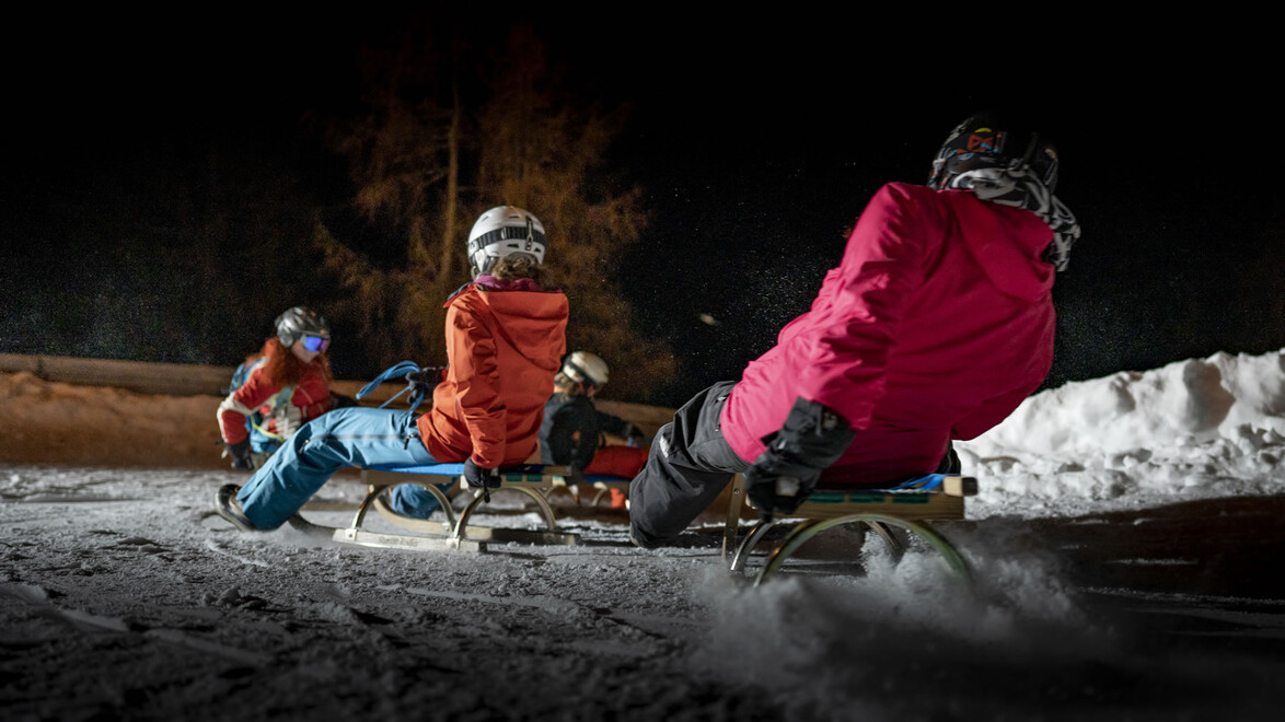 Person in pinker Jacke rodelt voran, zwei weitere folgen bei Nacht auf einer beleuchteten Bahn / A person in a pink jacket leads while two others follow on a lit toboggan track at night