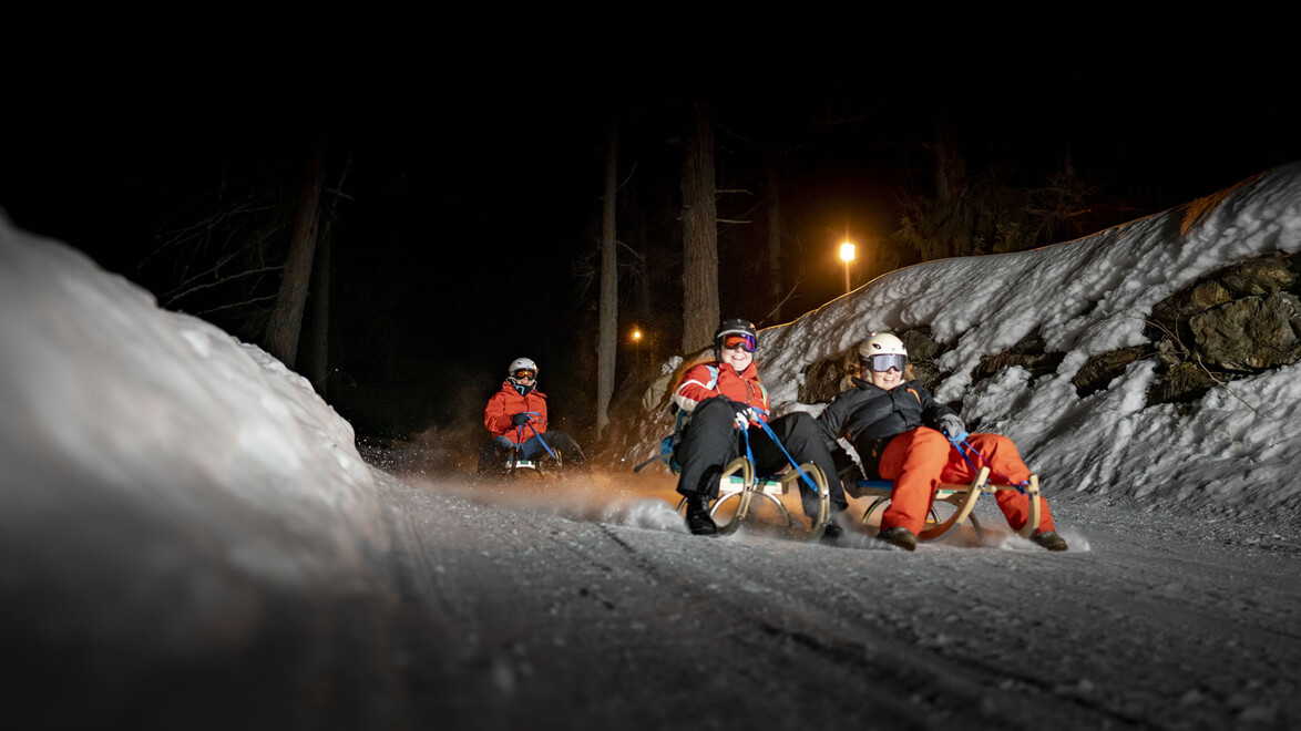 Drei Rodler:innen fahren bei Nacht mit Stirnlampen durch eine Kurve einer Rodelstrecke im Wald / Three sledders with headlamps navigate a turn of a forest toboggan trail at night