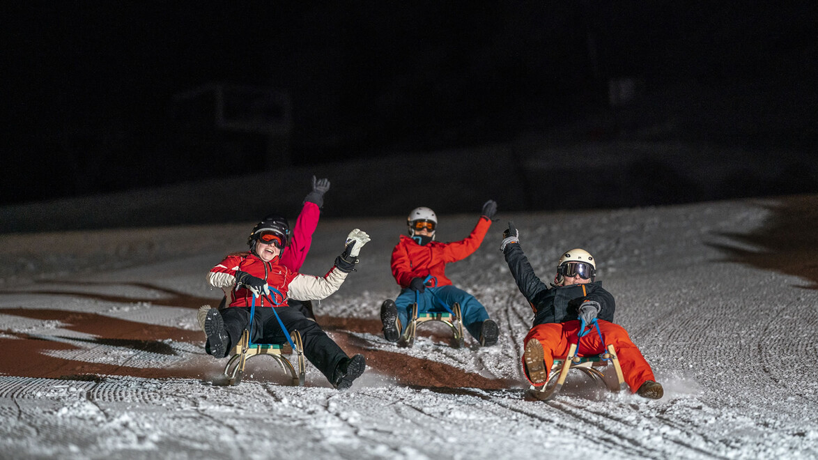Drei Rodler:innen fahren bei Nacht mit Stirnlampen eine beleuchtete Rodelbahn hinab / Three tobogganers ride down a lit toboggan run at night with headlamps