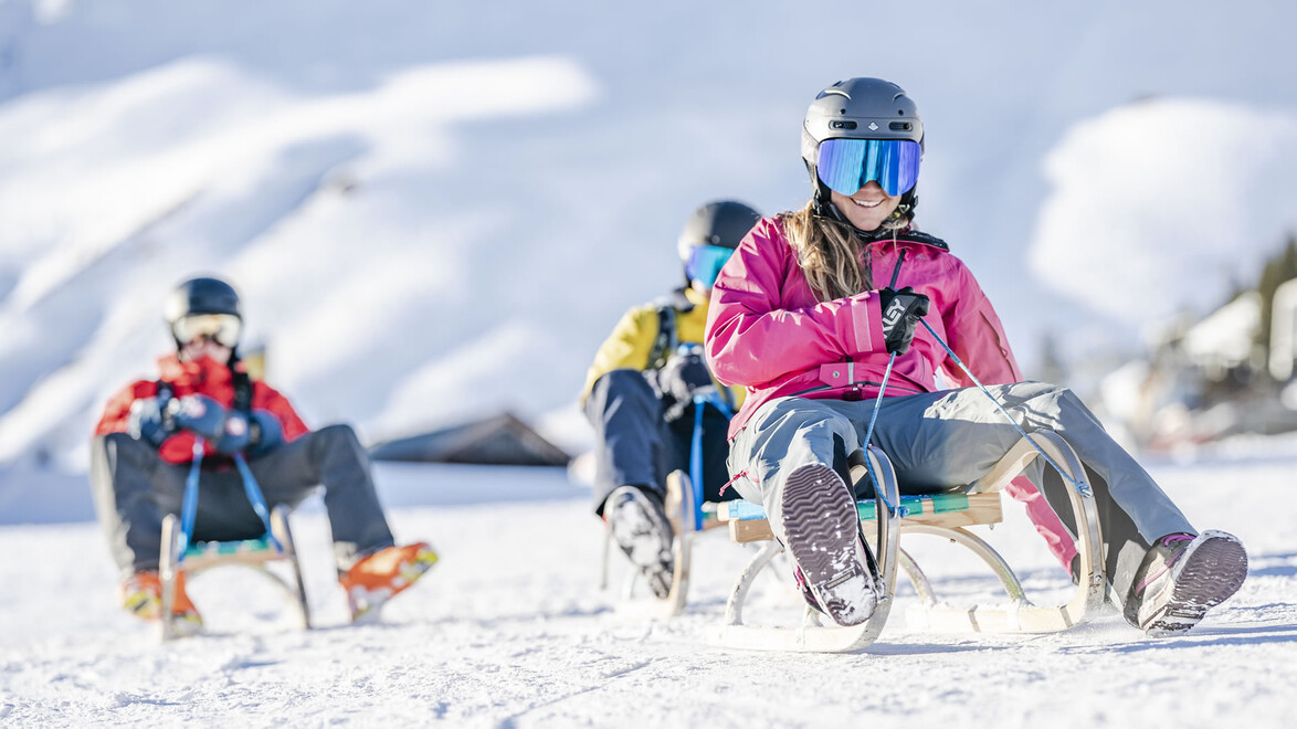 Vorneweg rodelt eine Person mit pinker Jacke, gefolgt von zwei weiteren im Schnee bei Tageslicht / A person in a pink jacket leads the toboggan run, followed by two others in the snow during daylight