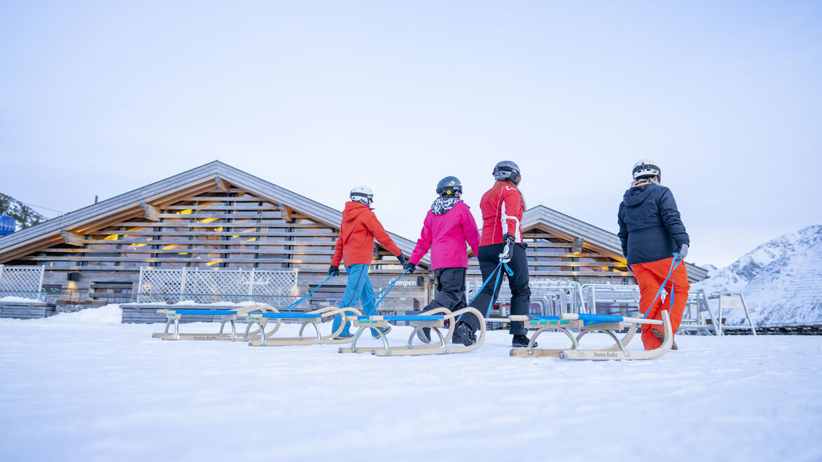 Vier Menschen mit Rodeln stehen vor einer verschneiten Berghütte / Four people with sleds stand in front of a snowy mountain hut