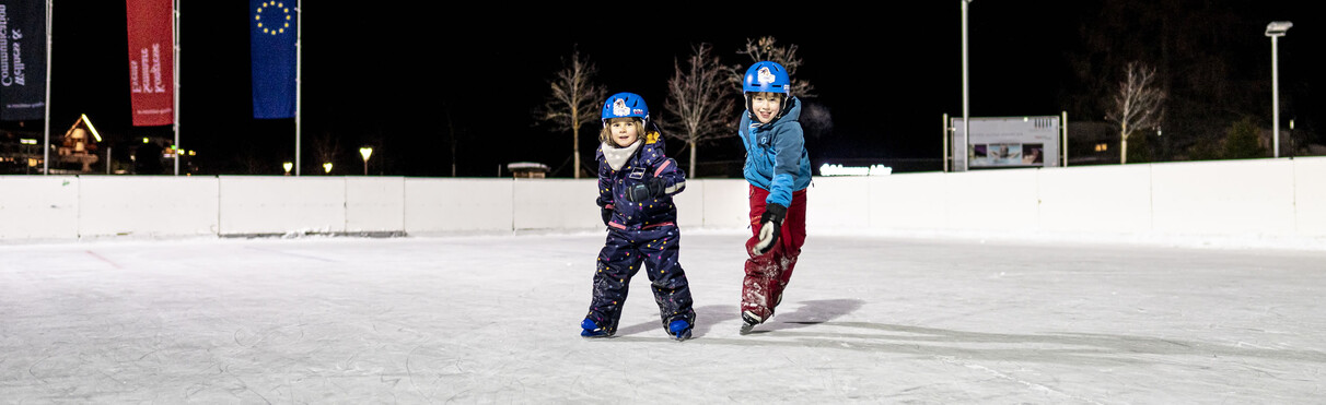 Two children with helmets skate on a lit ice rink at night.
