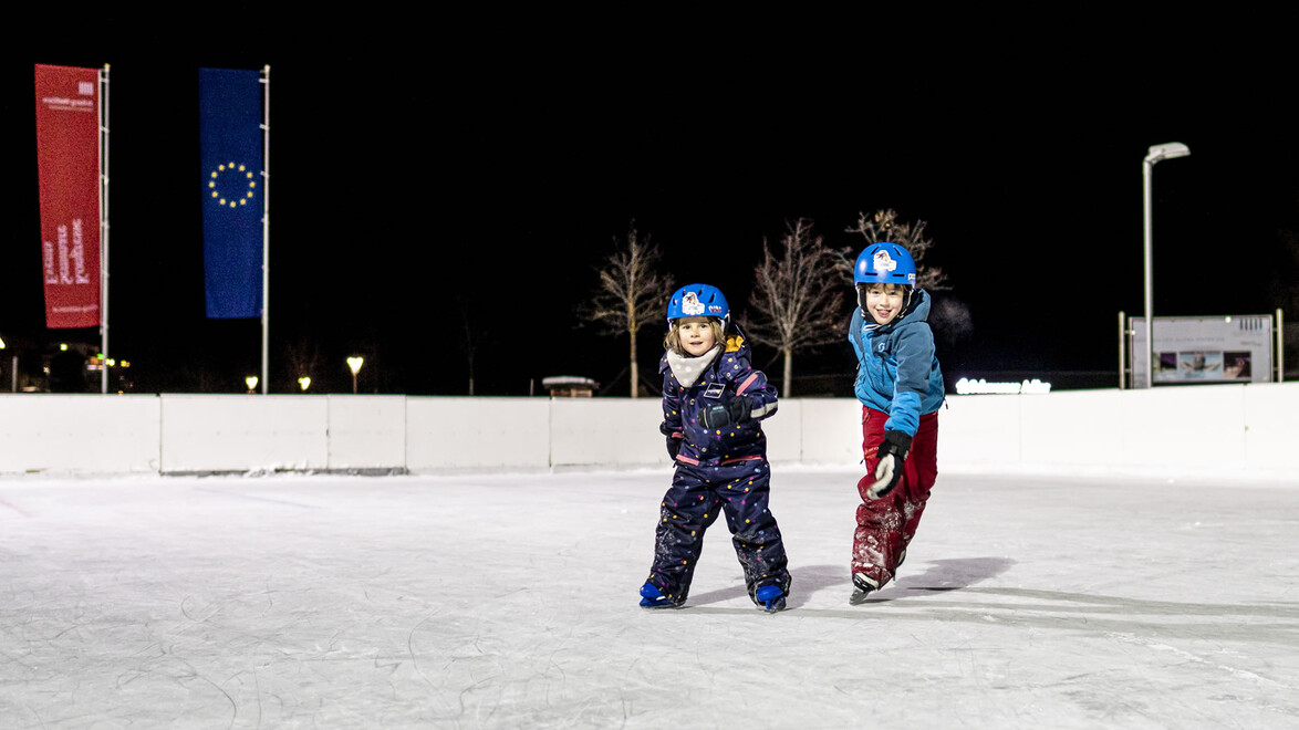 Zwei Kinder in Schneebekleidung gleiten auf dem Eis in Richtung Kamera unter freiem Himmel / Two kids in winter gear skate across the ice under the open sky