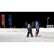 Zwei Kinder in Schneebekleidung gleiten auf dem Eis in Richtung Kamera unter freiem Himmel / Two kids in winter gear skate across the ice under the open sky