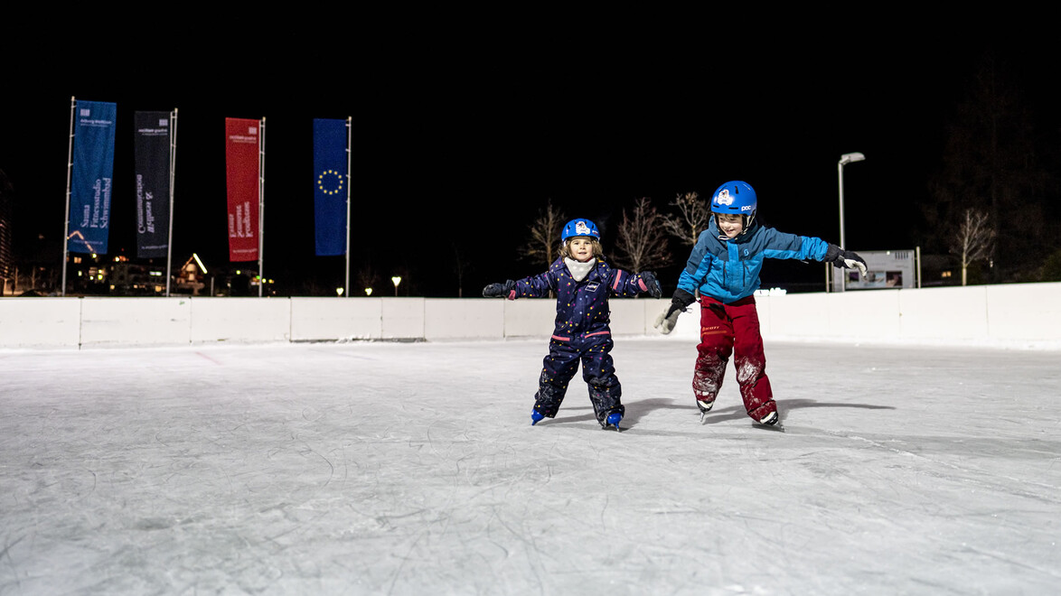 Zwei Kinder mit Helmen üben am Abend gemeinsam das Eislaufen auf einer beleuchteten Eisfläche / Two helmeted children practice skating together in the evening on a lit ice rink
