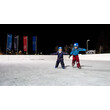 Zwei Kinder mit Helmen üben am Abend gemeinsam das Eislaufen auf einer beleuchteten Eisfläche / Two helmeted children practice skating together in the evening on a lit ice rink