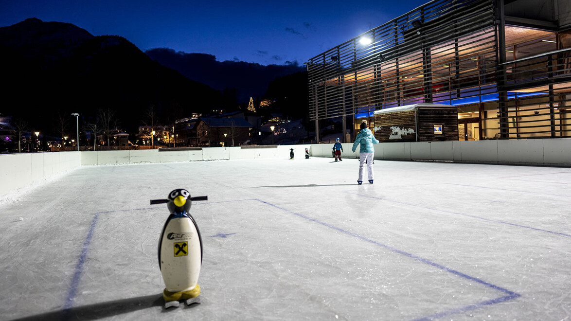Kinder lernen mit Pinguin-Lernhilfen auf einer abendlichen Kunsteisbahn das Schlittschuhlaufen / Children learn to skate with penguin aids on an evening ice rink