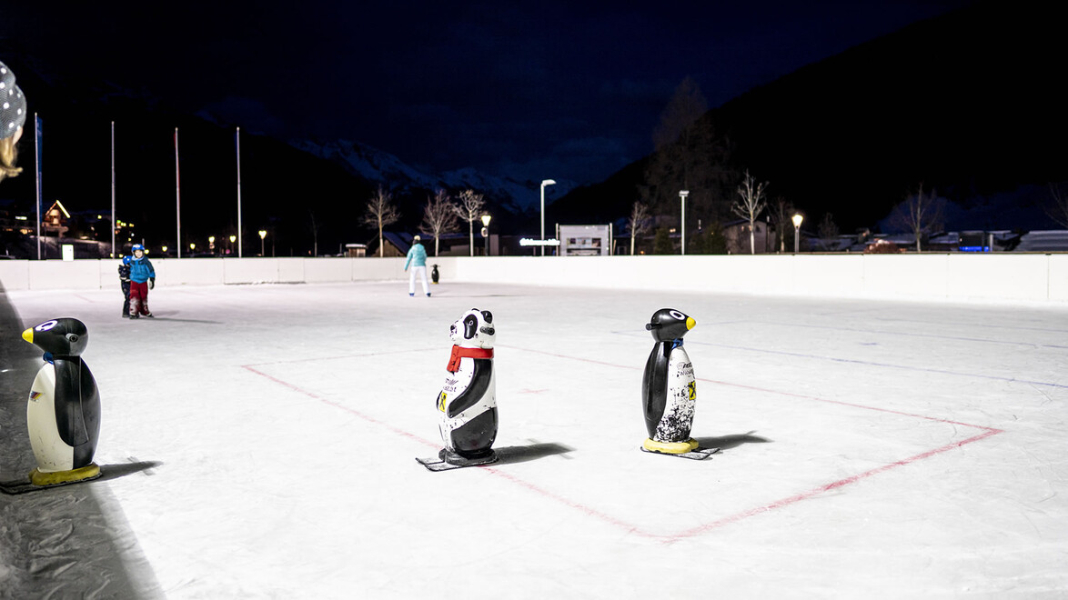 Mehrere Pinguinfiguren stehen am Rand einer nächtlich beleuchteten Kunsteisfläche bereit / Several penguin figures stand ready at the edge of a night-lit artificial ice rink