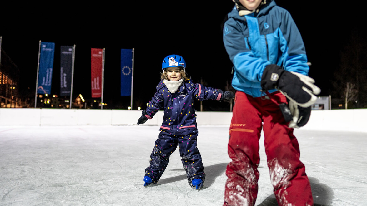 Nahaufnahme von zwei Kindern beim Eislaufen, begleitet von hellem Flutlicht und Fahnen im Hintergrund / Close-up of two kids ice skating under floodlights with flags in the background