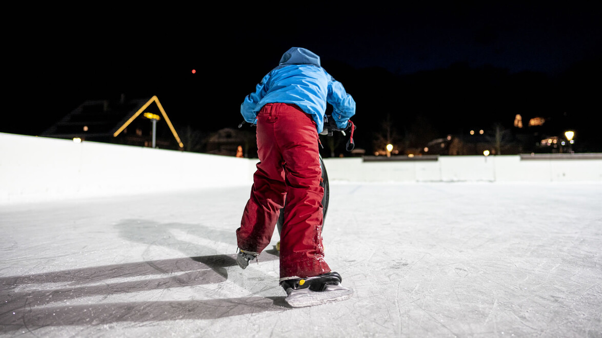 Ein Kind in roter Hose fährt am beleuchteten Eislaufplatz vorwärts, Stadtlichter im Hintergrund / A child in red pants skates forward on a lit rink with town lights behind