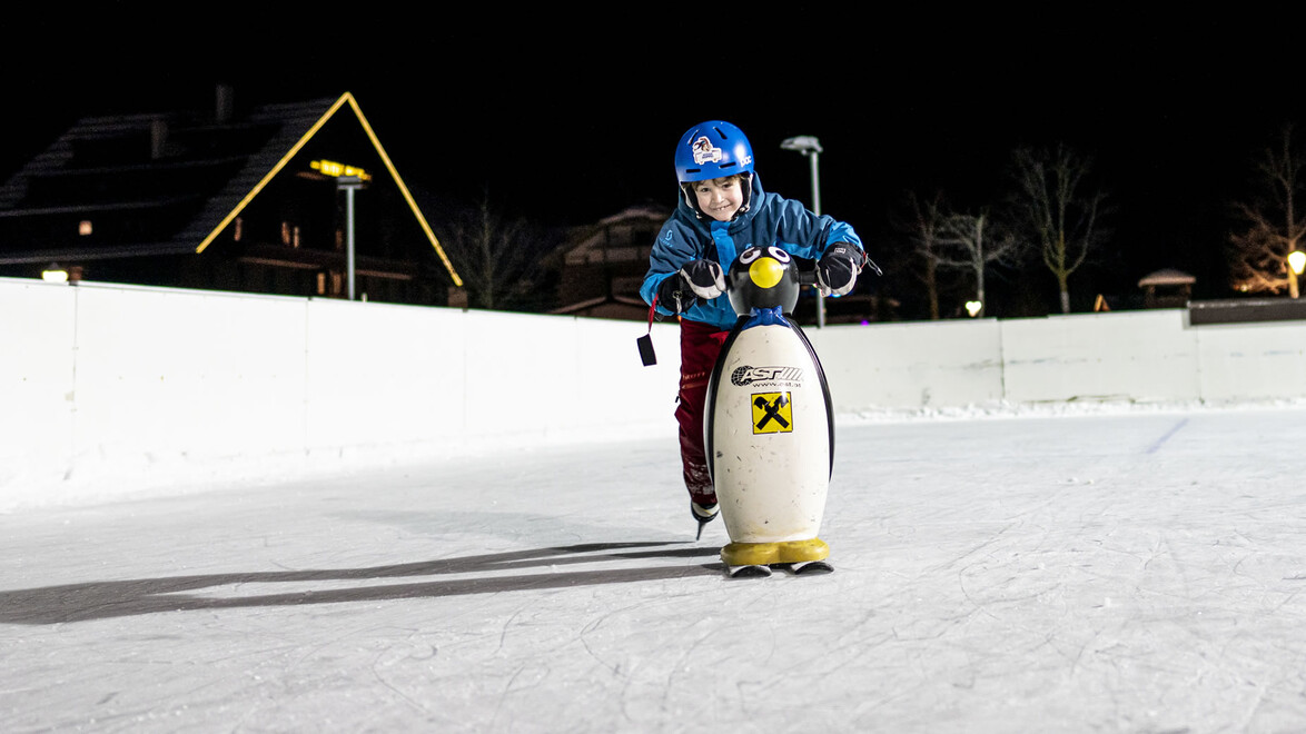 Ein Kind mit Helm hält sich auf der Eisfläche an einem Pinguin fest, um das Gleichgewicht zu üben / A helmeted child uses a penguin skating aid to practice balance on the ice