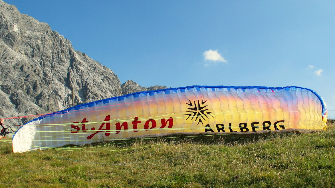 Paragleitschirm mit St. Anton-Schriftzug auf grüner Wiese vor Bergkulisse / Paragliding canopy with St. Anton lettering on meadow in front of mountain backdrop