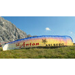 Paragleitschirm mit St. Anton-Schriftzug auf grüner Wiese vor Bergkulisse / Paragliding canopy with St. Anton lettering on meadow in front of mountain backdrop