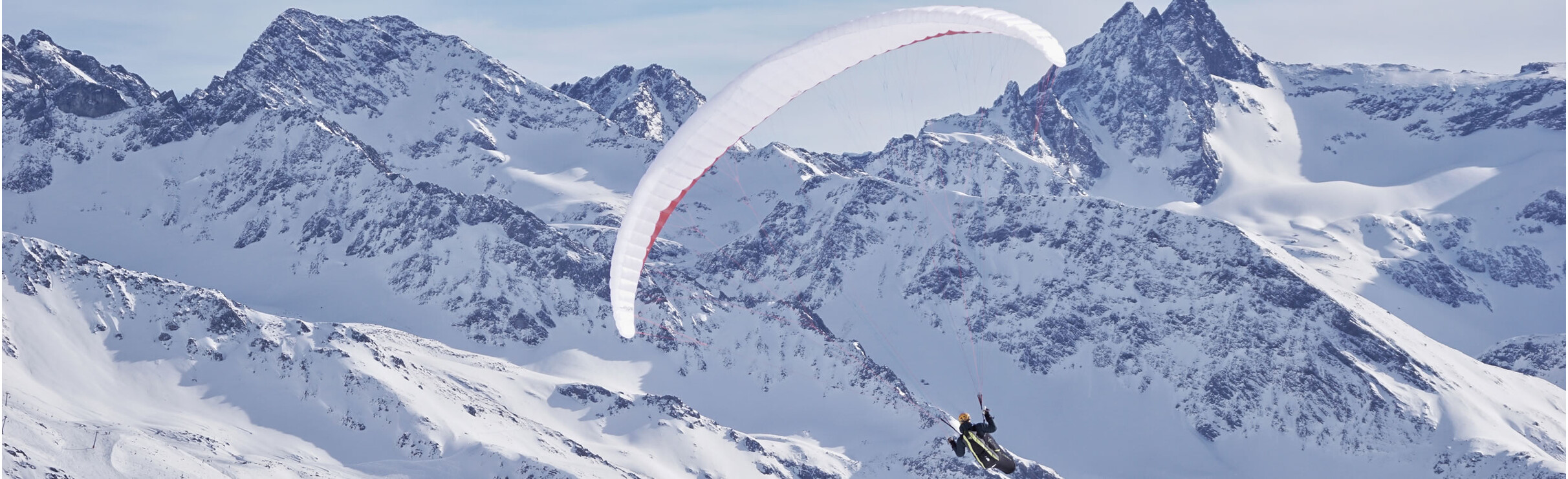 A paraglider flies over a snow-covered alpine landscape with striking mountain peaks in the background.