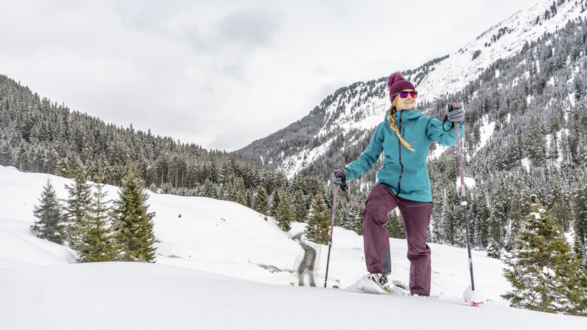 Frau mit Schneeschuhen steht auf einem Hang und blickt in das winterliche Tal / Woman with snowshoes stands on a slope and looks into the wintry valley