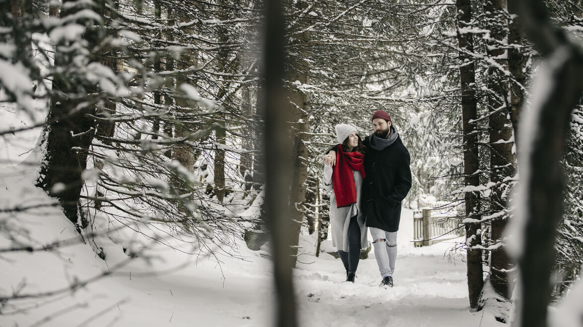 Paar spaziert durch einen verschneiten, ruhigen Waldpfad im Winter / Couple walks through a snow-covered, peaceful forest trail in winter