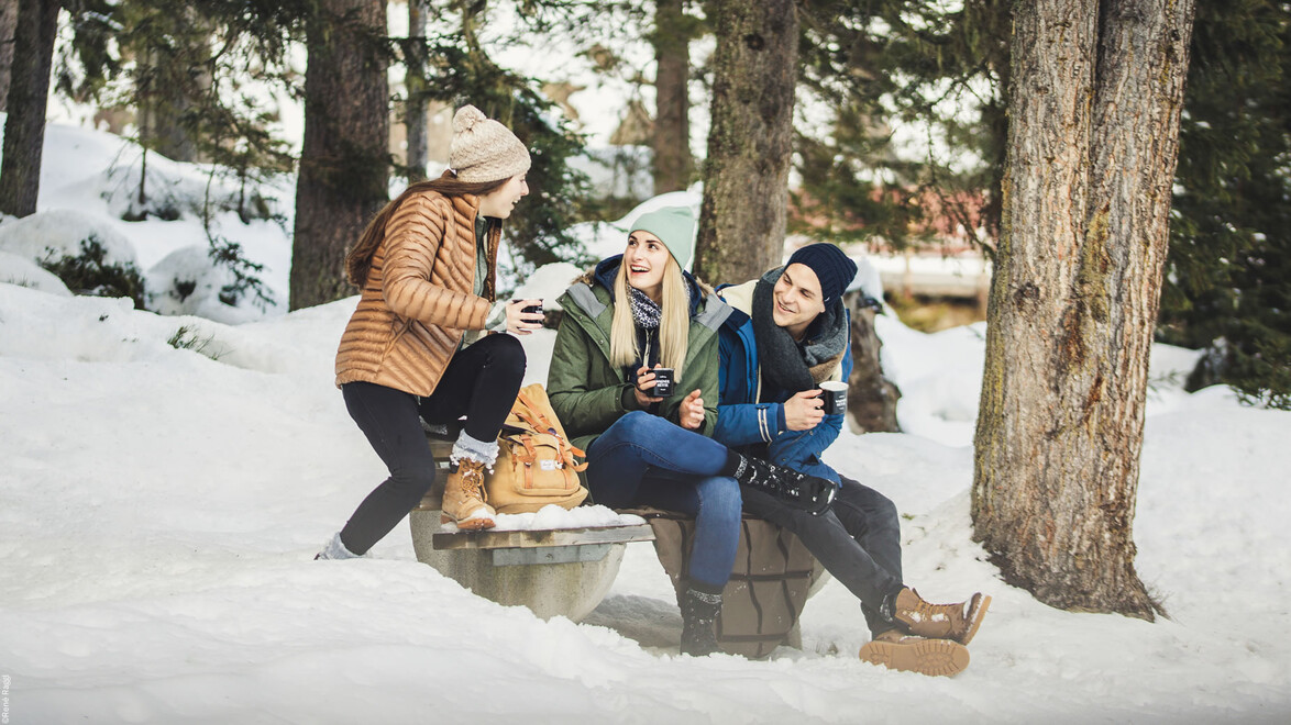 Drei Frauen sitzen lachend auf einem Baumstamm im verschneiten Winterwald / Three women sit laughing on a tree trunk in the snowy winter forest
