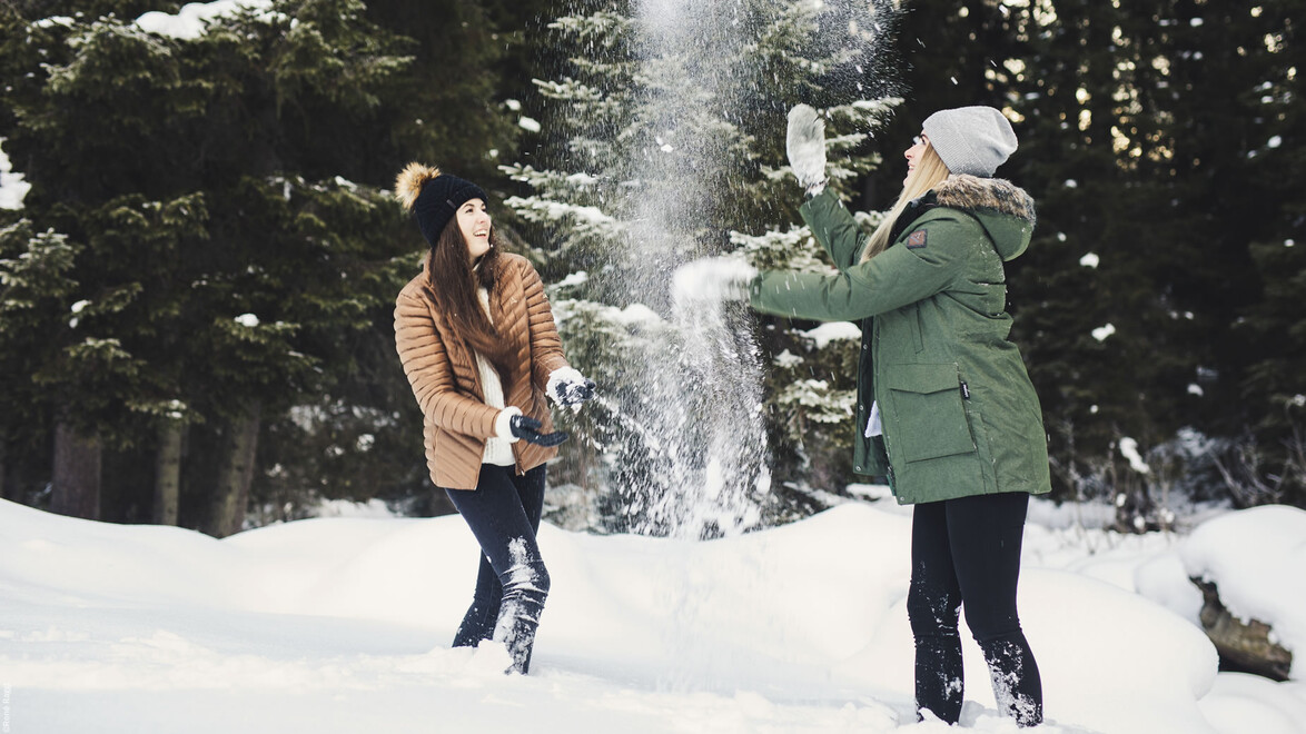 Zwei Frauen spielen lachend im Schnee zwischen verschneiten Bäumen / Two women play joyfully in the snow between snowy trees