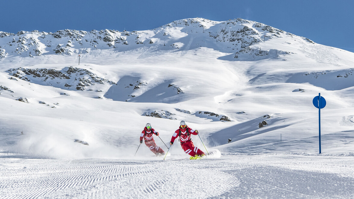Zwei Skifahrer:innen fahren auf einer breiten Piste mit weichem Pulverschnee vor eindrucksvoller Gebirgskulisse / Two skiers descend a wide slope with soft powder snow and impressive mountain backdrop