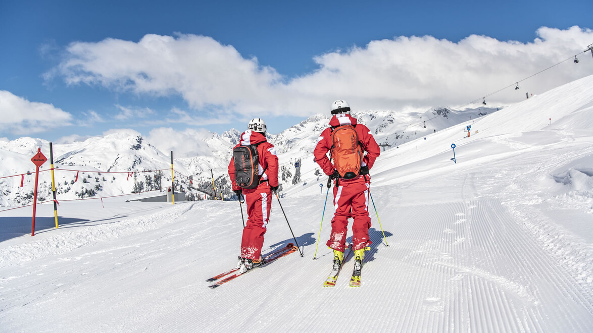 Zwei Skiführer in roter Bekleidung stehen auf einer präparierten Piste mit Alpenpanorama / Two ski guides in red outfits stand on a groomed slope with alpine panorama