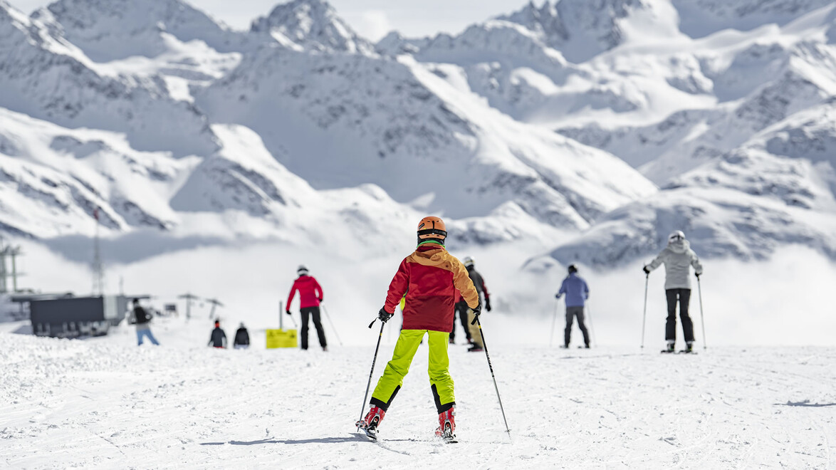 Kinder und Erwachsene fahren auf einer Skipiste, die von hohen Bergen umgeben ist / Children and adults ski on a slope surrounded by high mountains
