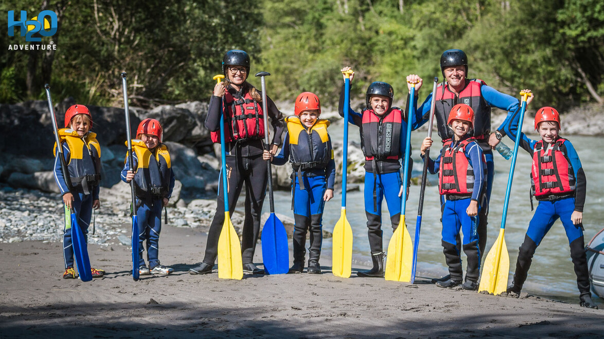 Gruppe mit Kindern und Erwachsenen in Neoprenanzügen mit Paddeln vor dem Rafting / Group of children and adults in wetsuits with paddles before rafting