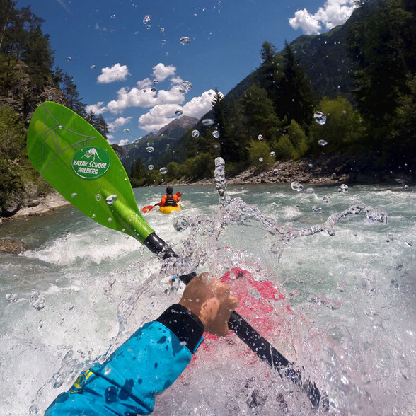 Kayaker paddles through splashing whitewater with scenic green mountains under a blue sky.