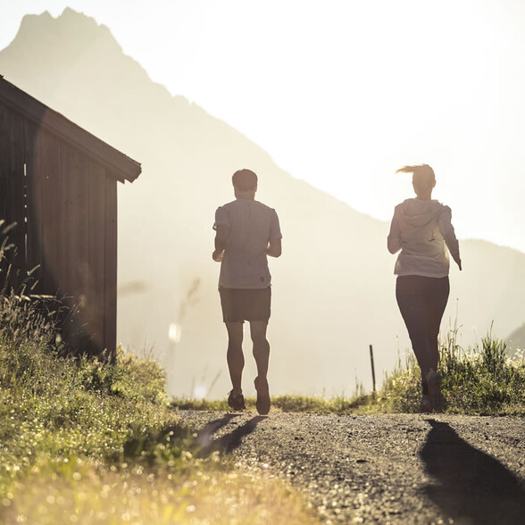 Zwei Jogger laufen bei Sonnenaufgang auf einem Pfad neben einer Holzhütte.