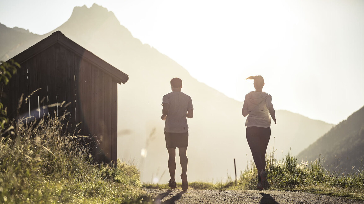 Zwei Menschen joggen in der Abendsonne einen Weg entlang, im Hintergrund ein Bergmassiv und eine Hütte / Two people jog along a path in the evening sun, with a mountain massif and a hut in the background